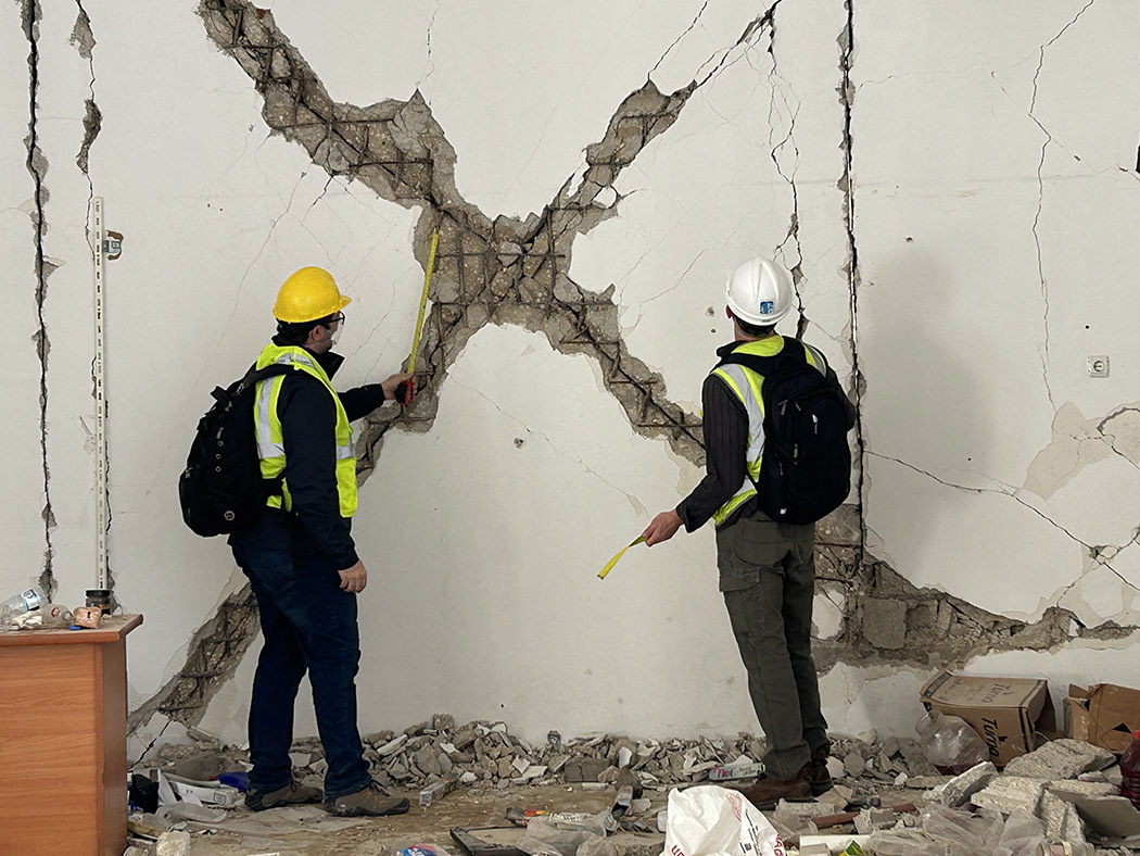 Two people wearing safety gear and backpacks and holding tape measures face away from the camera toward a wall with an X-shaped damaged area. 