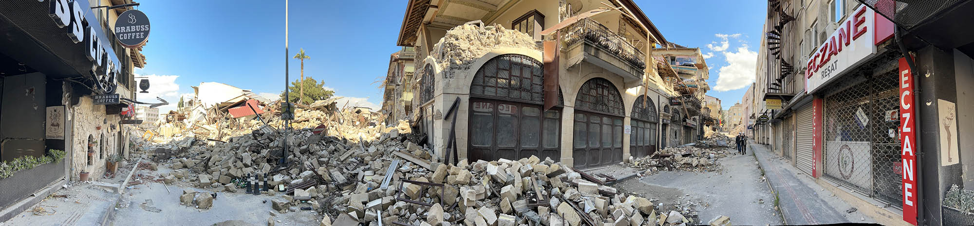 Wide-angle photo shows two rubble-filled streets lined with damaged buildings that once housed coffeeshops and other businesses.