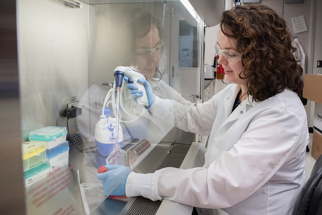 Jerilyn Izac sits at a lab bench with her hands inside a plastic shield, using a device to draw liquid from a plastic container into a tube. 