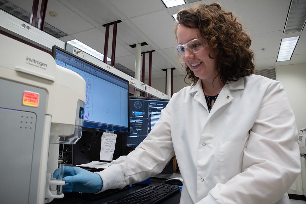 Jerilyn Izac, wearing safety glasses and a lab coat, places a test tube into a holder on a large piece of lab equipment.