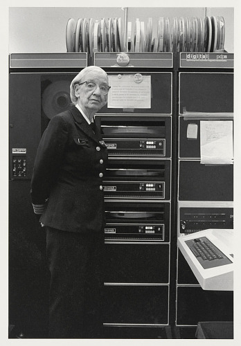 Historical photo shows Grace Hopper in uniform standing next to a rack of computer equipment.