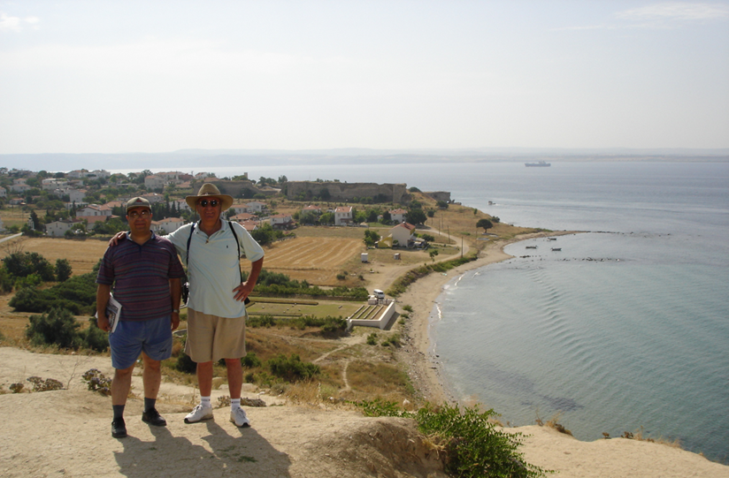 Two men in hats and sunglasses pose standing on a cliff over the beach. 