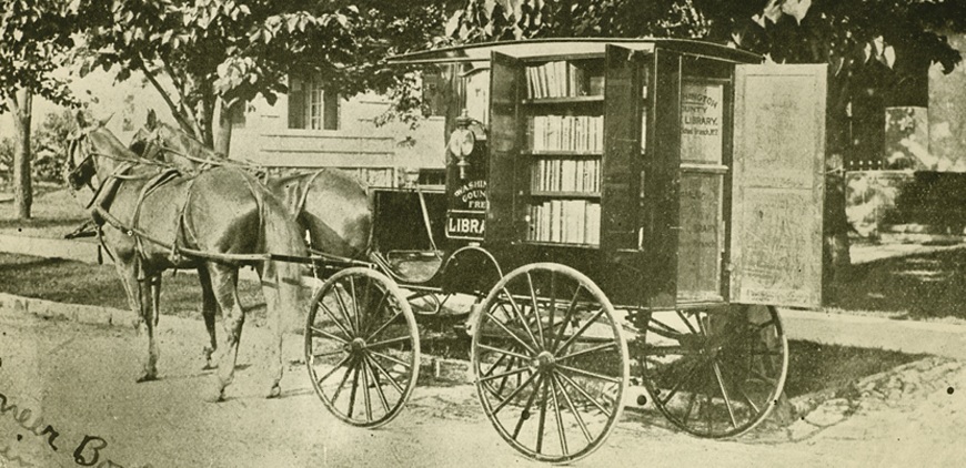 Historical photo shows two horses harnessed to a wagon with open doors on the outside showing shelves holding books. 