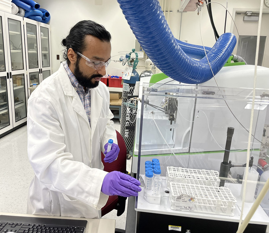 George Caceres wears safety glasses and a lab coat as he places a sample in a test tube inside clear plastic box holding scientific machinery.