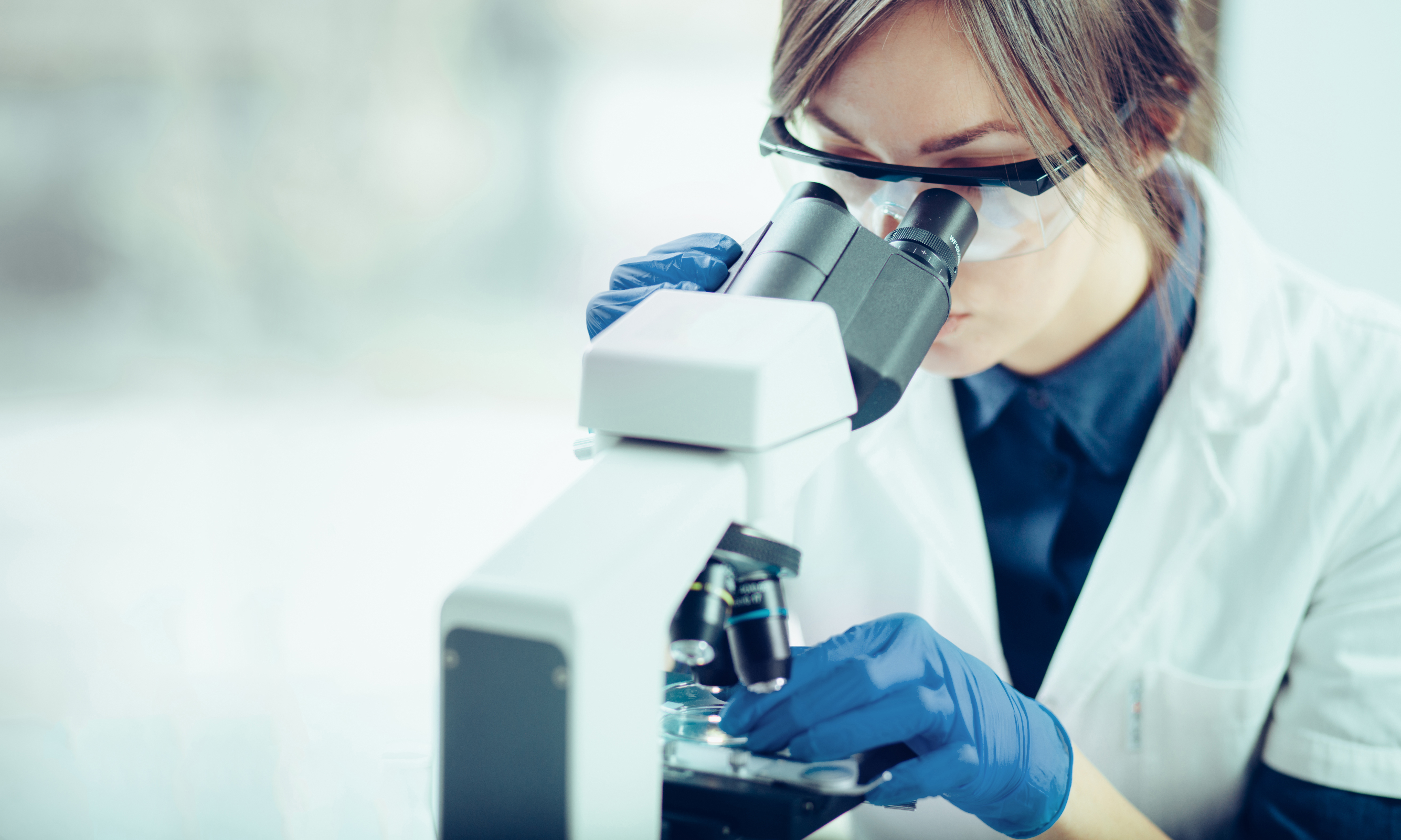 A young female researcher wearing safety glasses and a lab coat looks into a microscope.