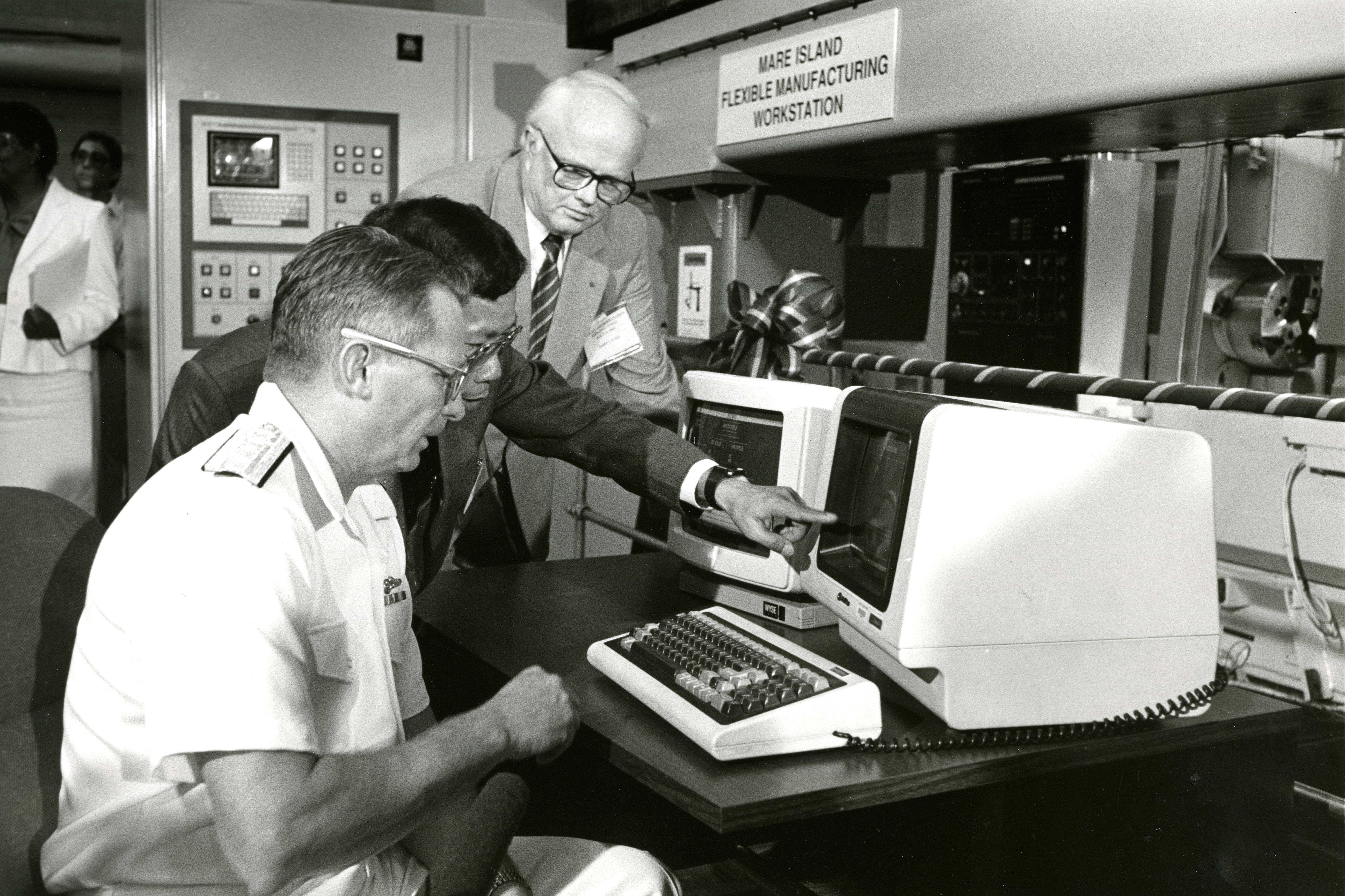 A black and white photograph of a naval officer seated at a computer workstation, another man pointing to the workstation, and a third man standing nearby.