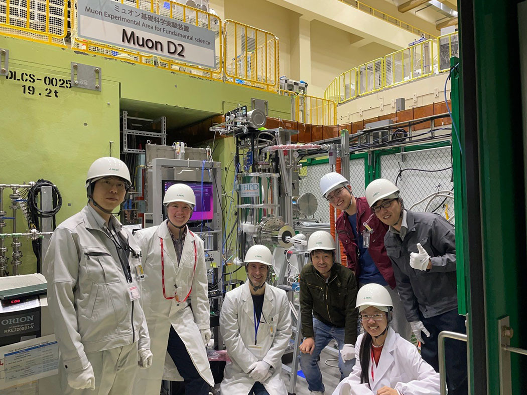 Seven researchers wearing white hard hats and lab coats pose for a group photo in front of large pieces of lab equipment. Sign above says: Muon D2.