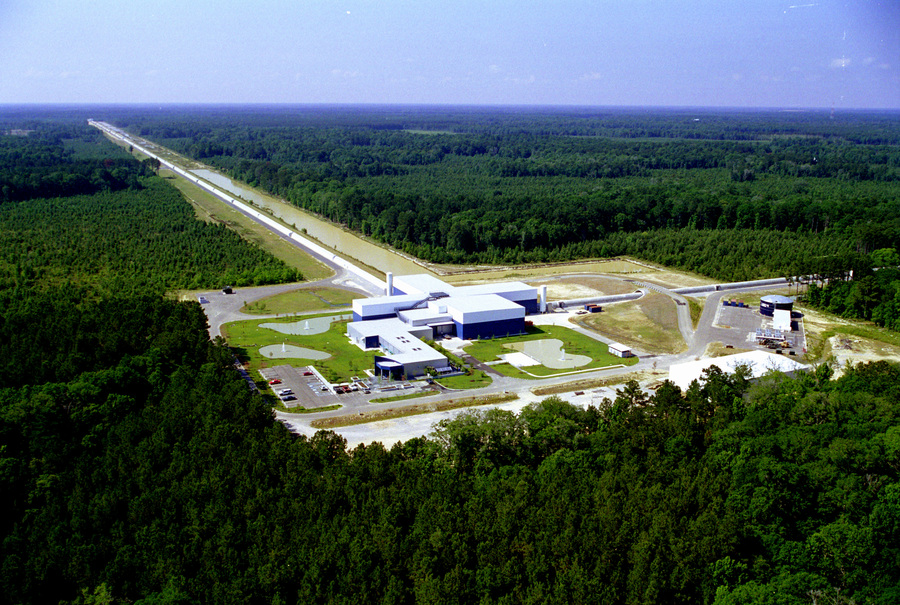 Aerial view of LIGO facility shows a white-roofed lab building surrounded by forest, with long thin paths extending from the building's back and side into the distance. 