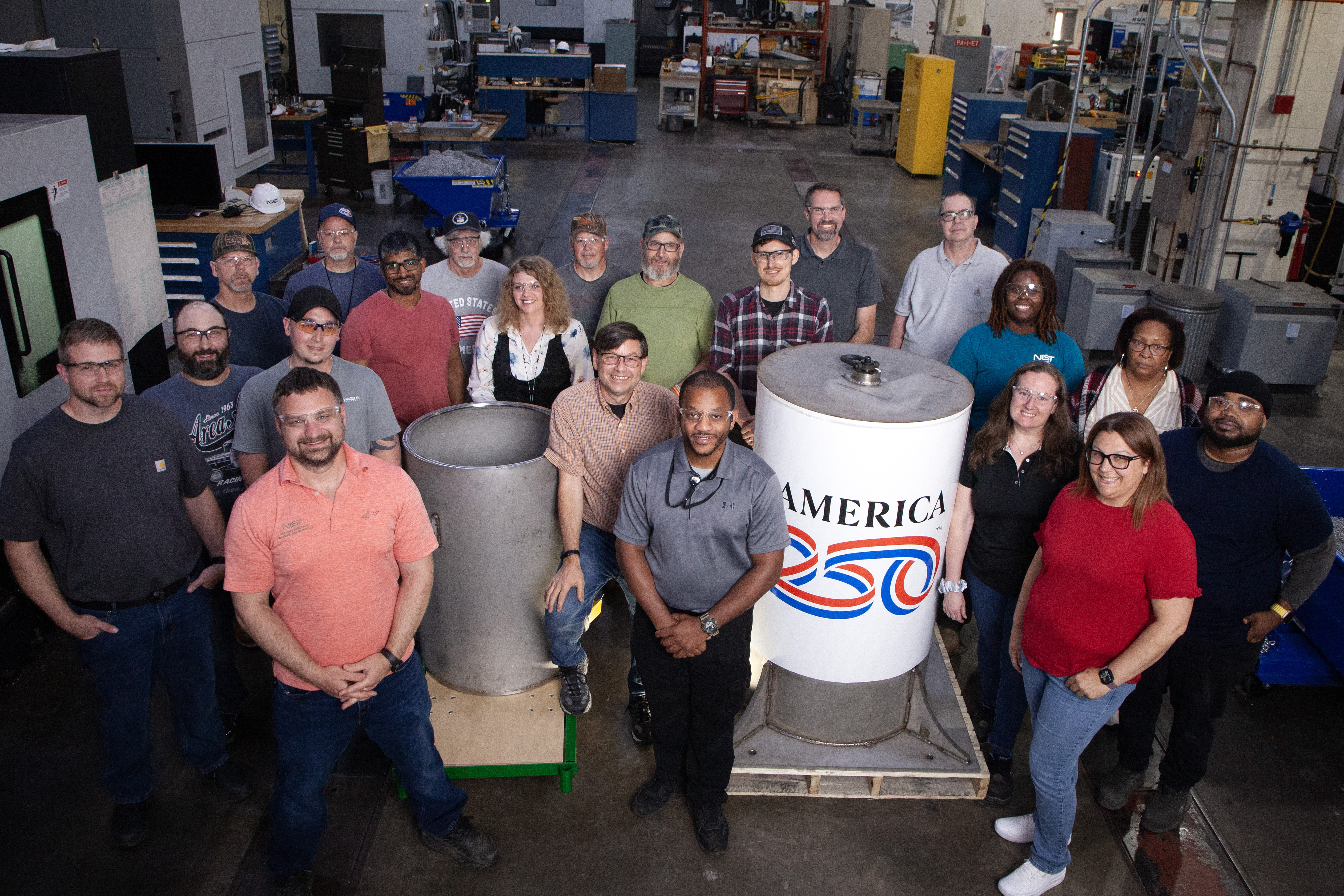 Almost two dozen people pose smiling in a large machine shop, standing around the cylindrical time capsule and the bell jar that will go over it, which is marked with "America 250."