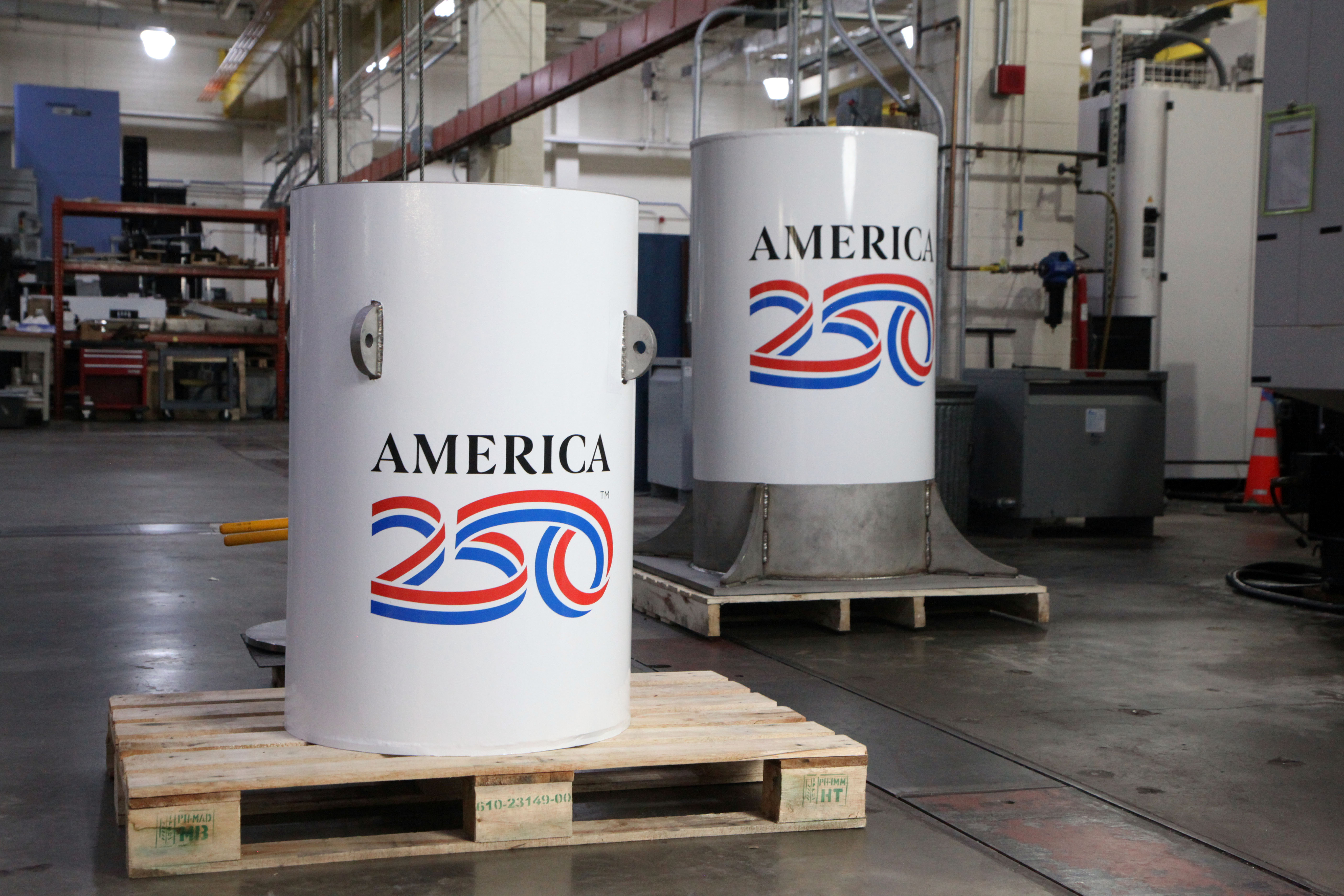 Two white cylinders with the America 250 logo stand in a large workshop space. 