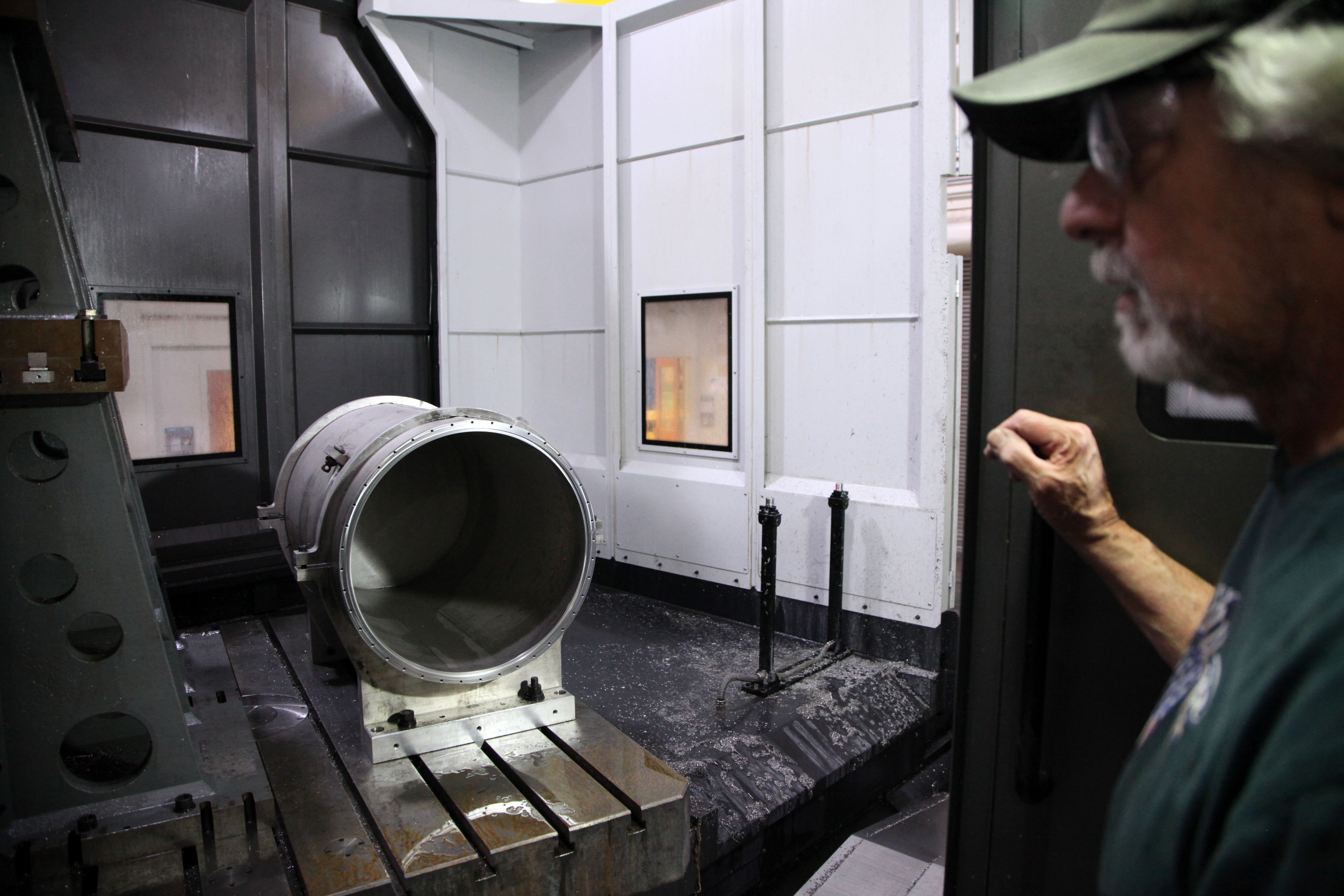 A man wearing safety glasses stands looking at a cylinder of thick metal lying on a wet platform inside a workshop. 