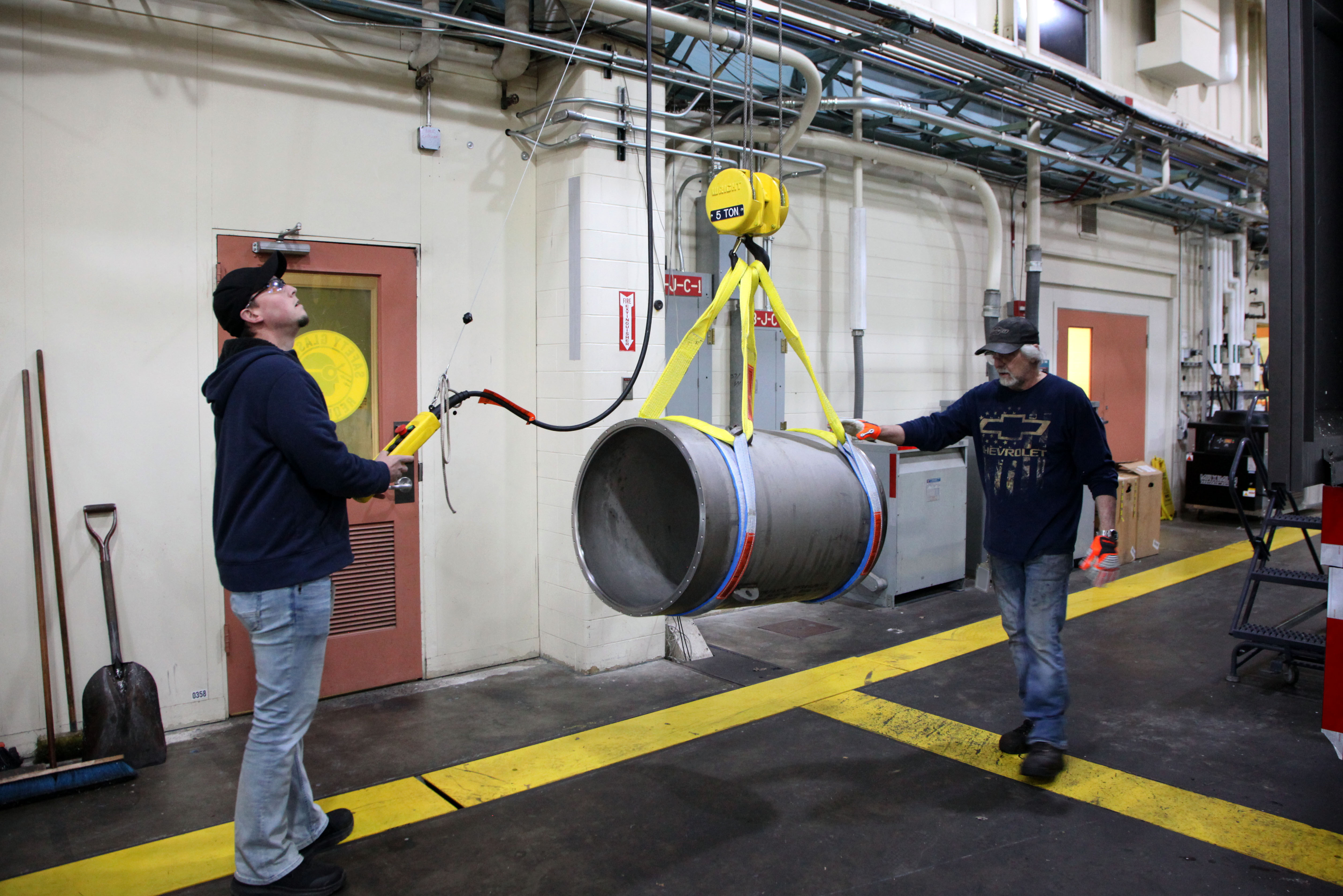Two men use an overhead pulley apparatus to move a thick metal cylinder inside a large workshop. 