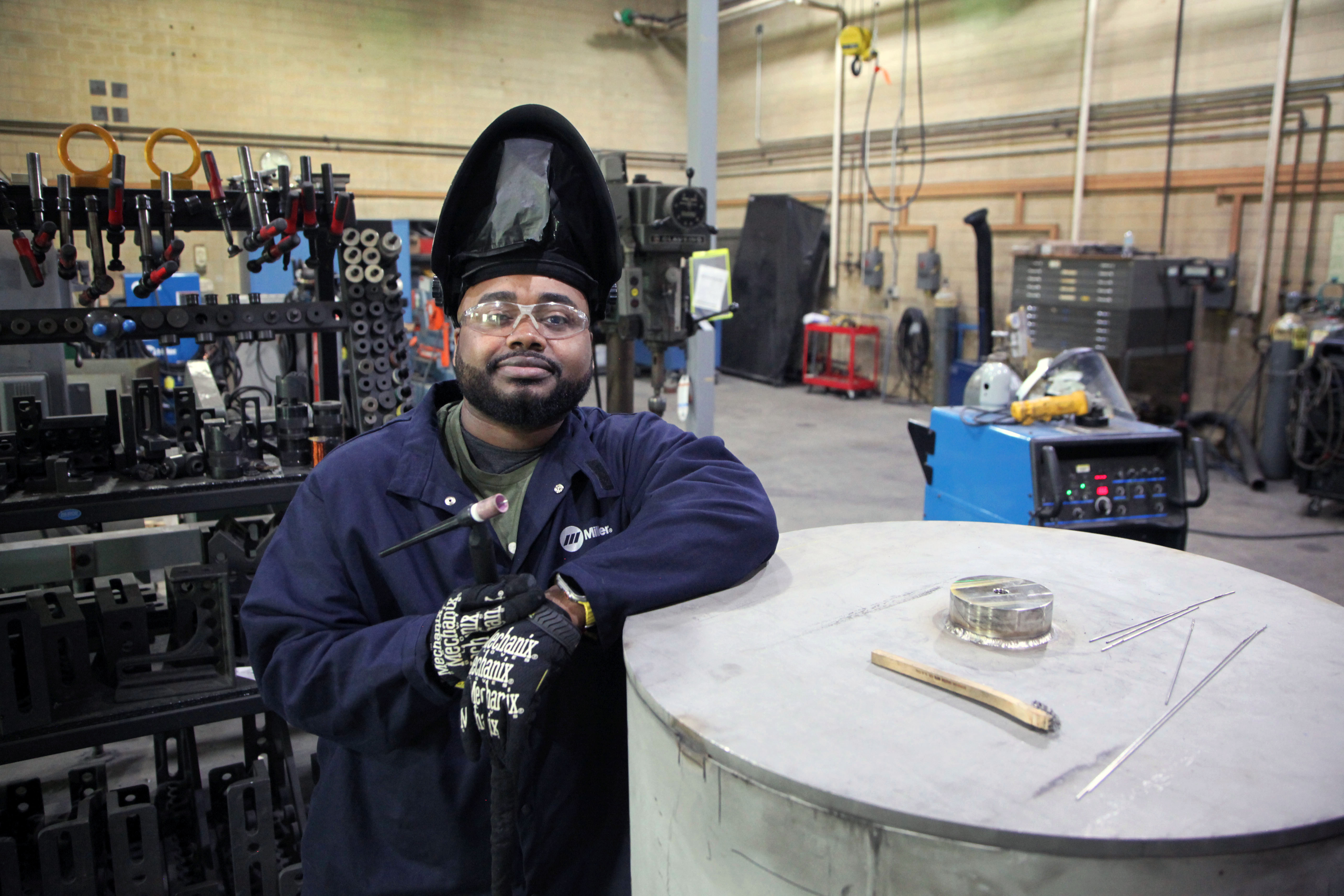A man wearing an uplifted welding mask and safety glasses poses leaning on a large metal cylinder in a workshop. 
