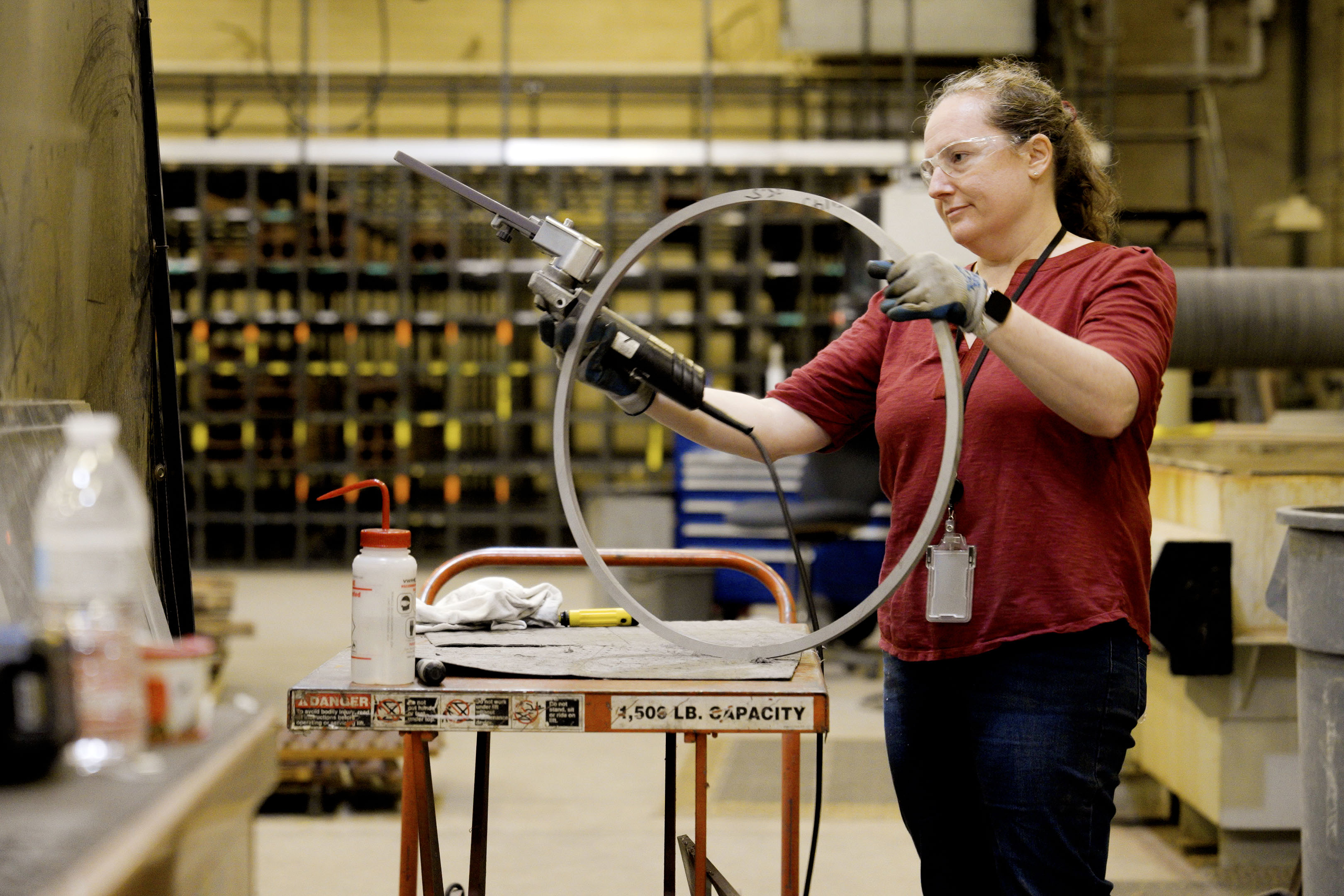 A woman wearing safety glasses holds an electric tool to work on a circle of metal in a large workshop. 