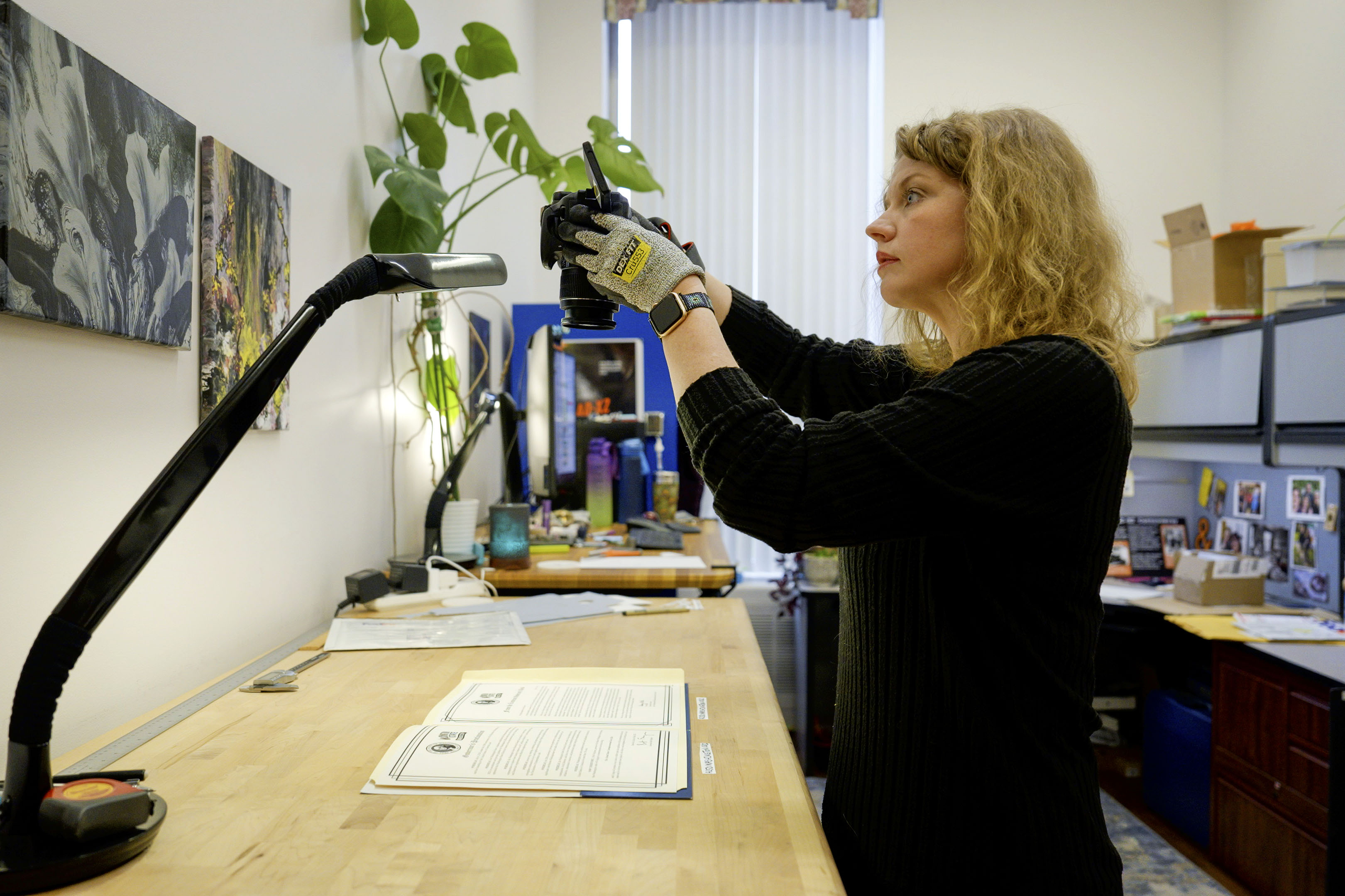A woman wearing gloves stands in an office holding a camera pointed down at papers on the desk.