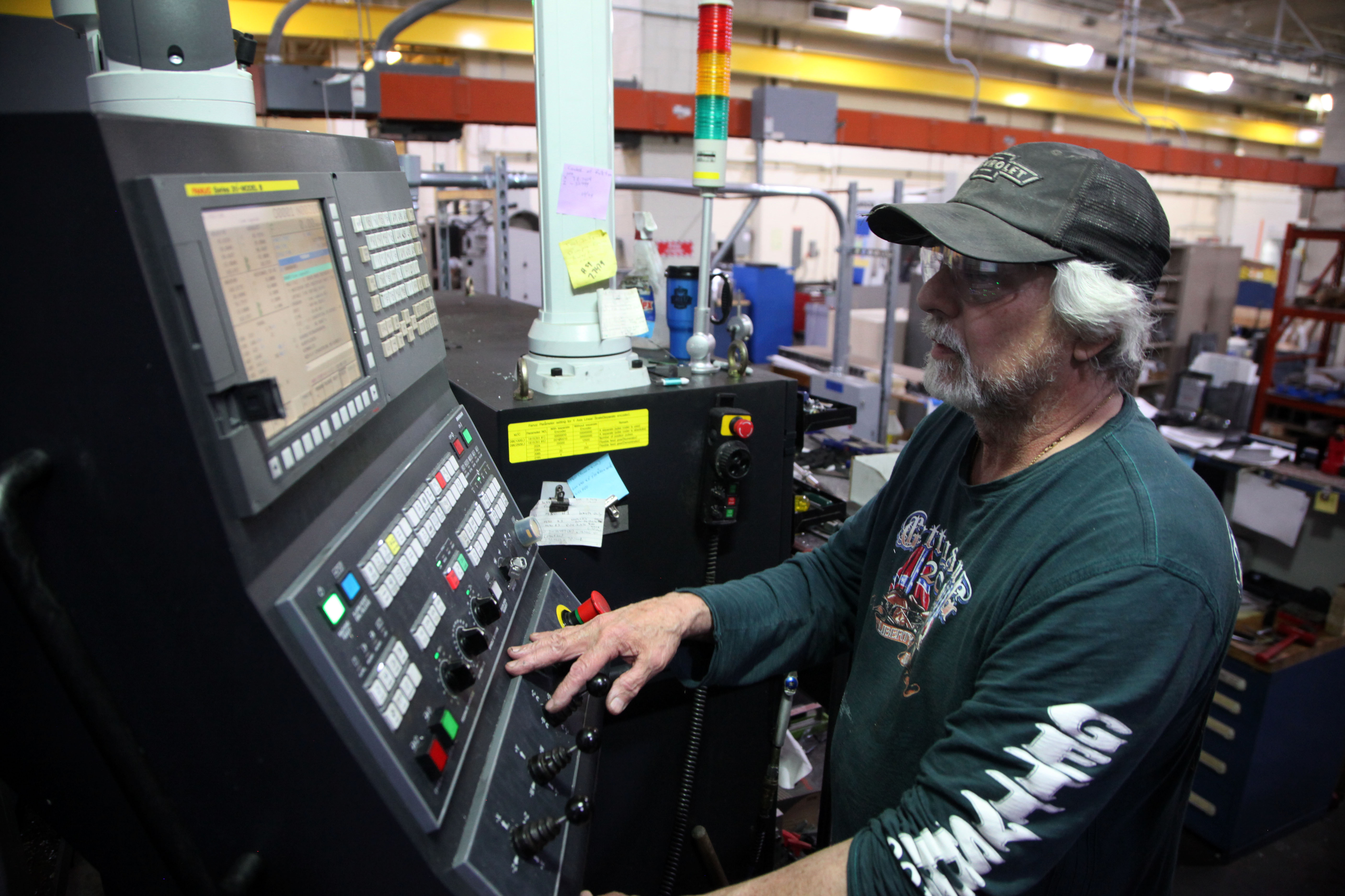 A man in a baseball cap stands at a control panel full of buttons inside a machine shop.
