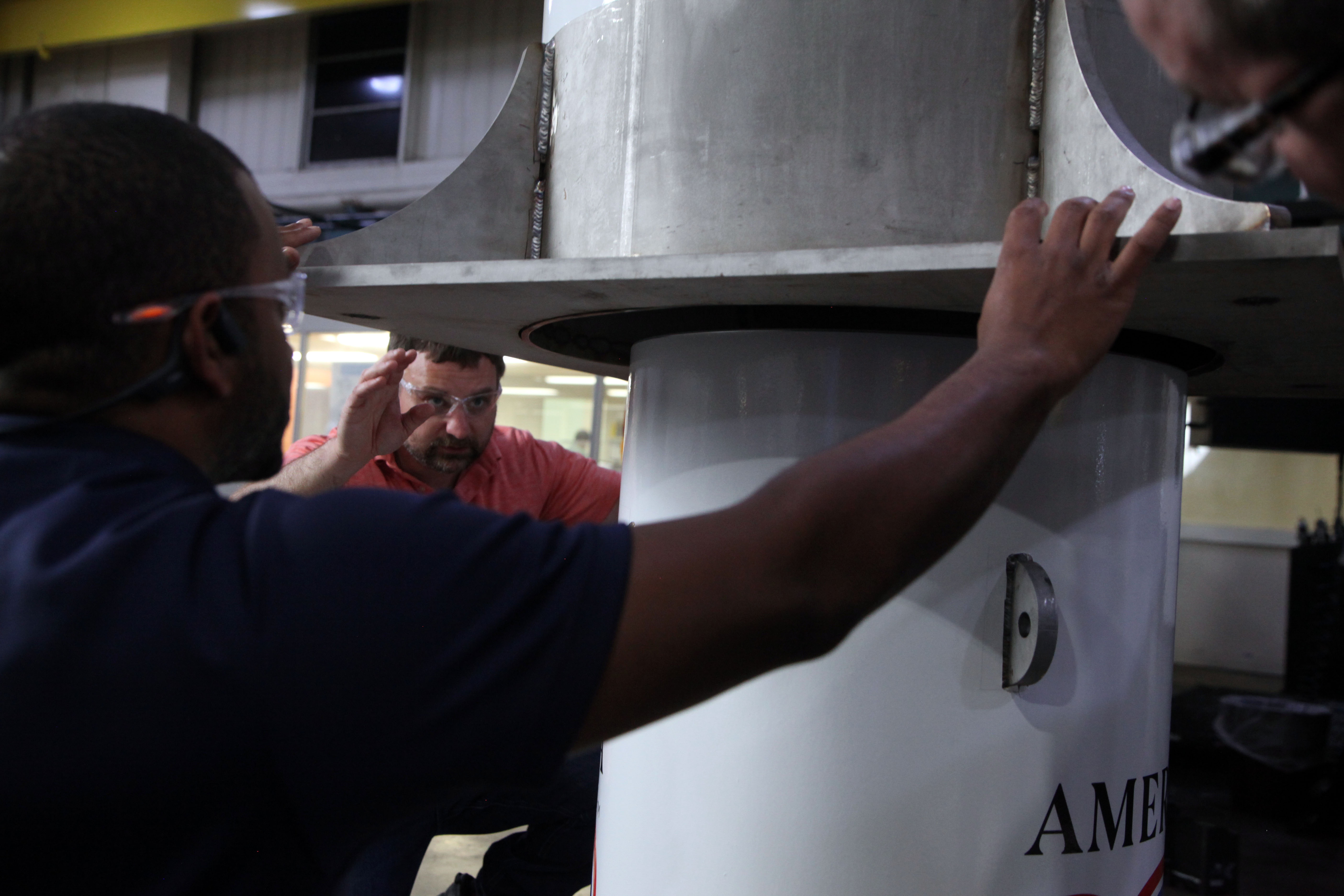 Two men reach out to hold a large metal cylinder being lowered onto a smaller one. 