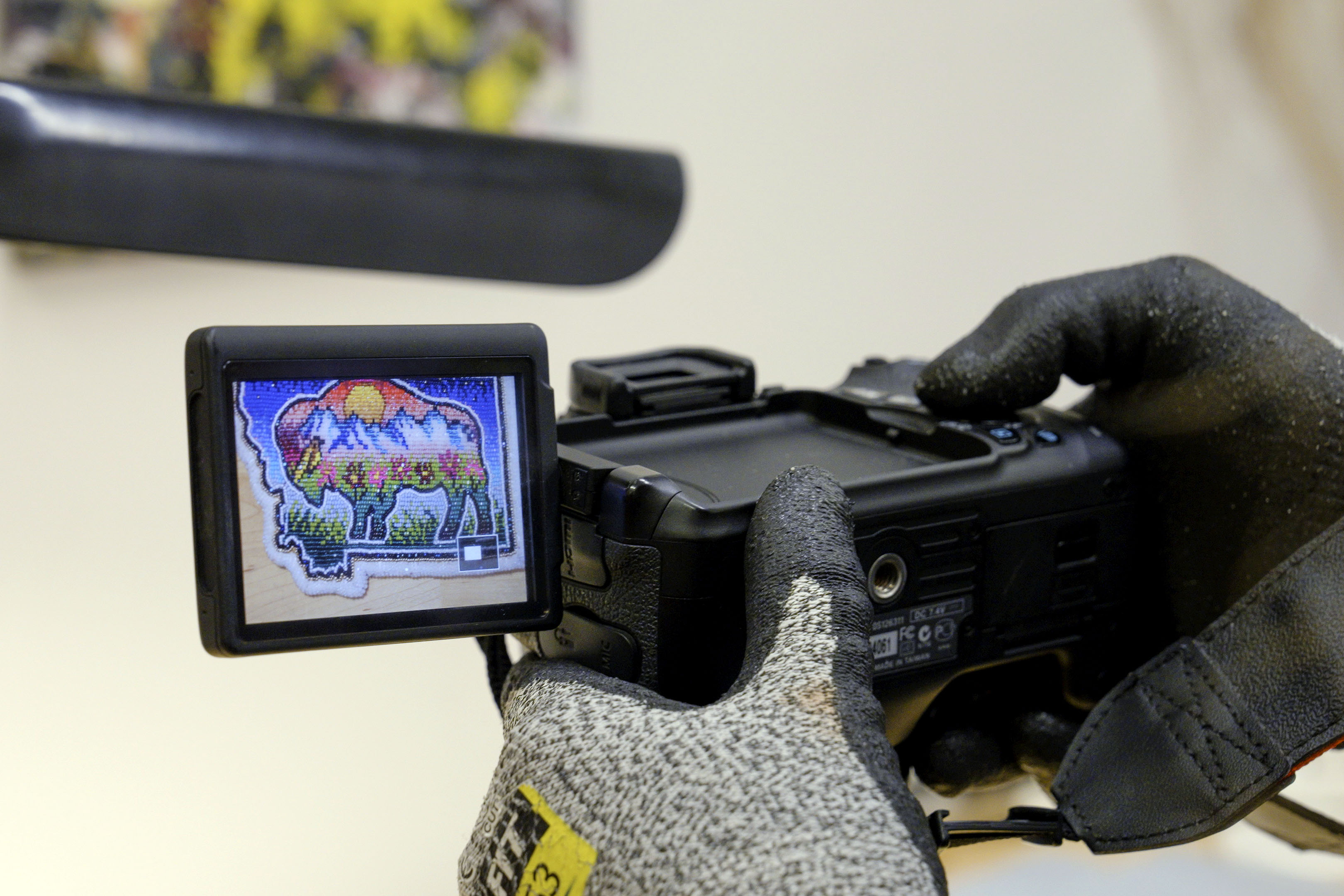 Gloved hands hold a camera; an attached screen shows a colorful beaded patch depicting a buffalo. 