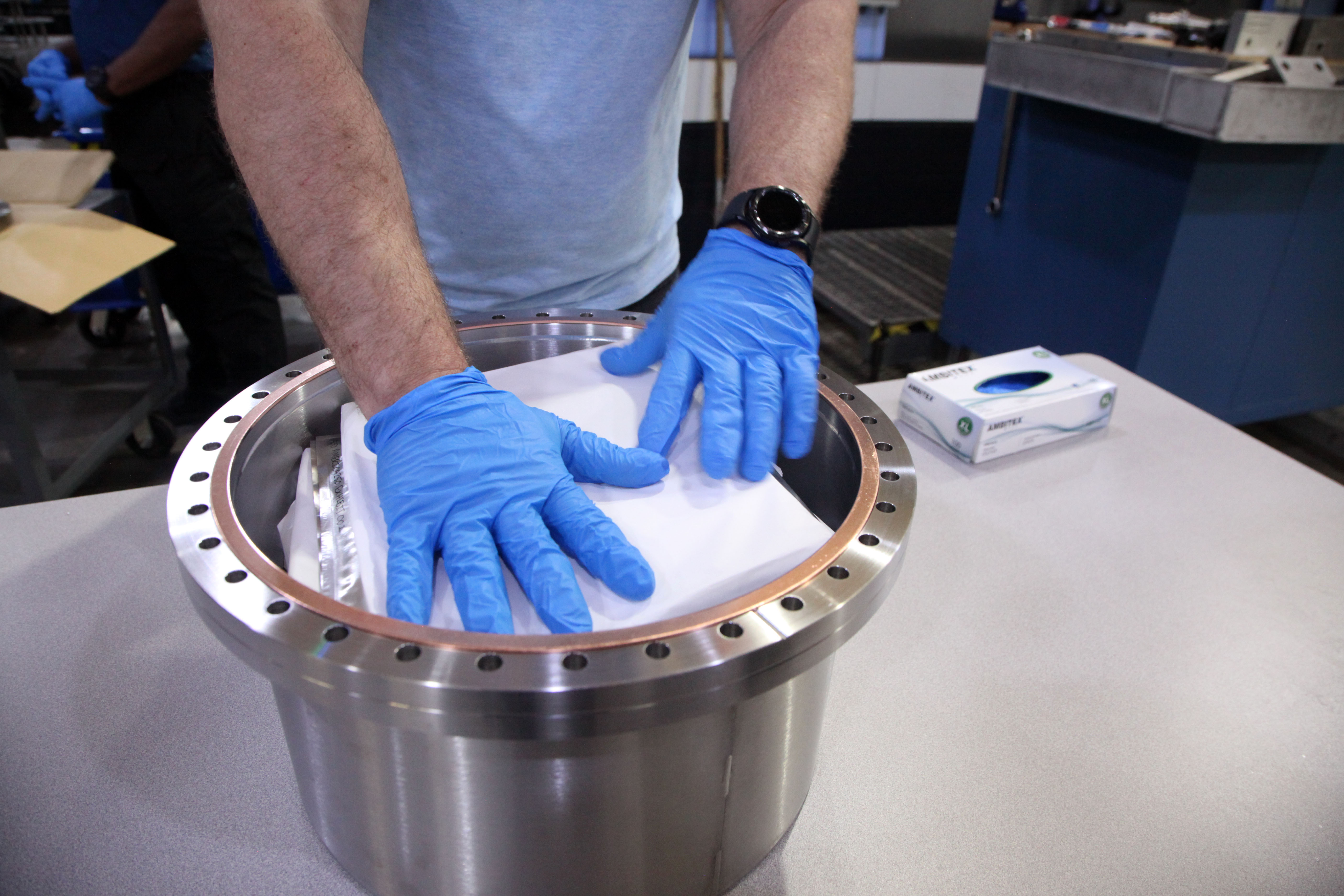 A person wearing blue gloves holds down folded paper on top of an open metal cylinder on a table. 