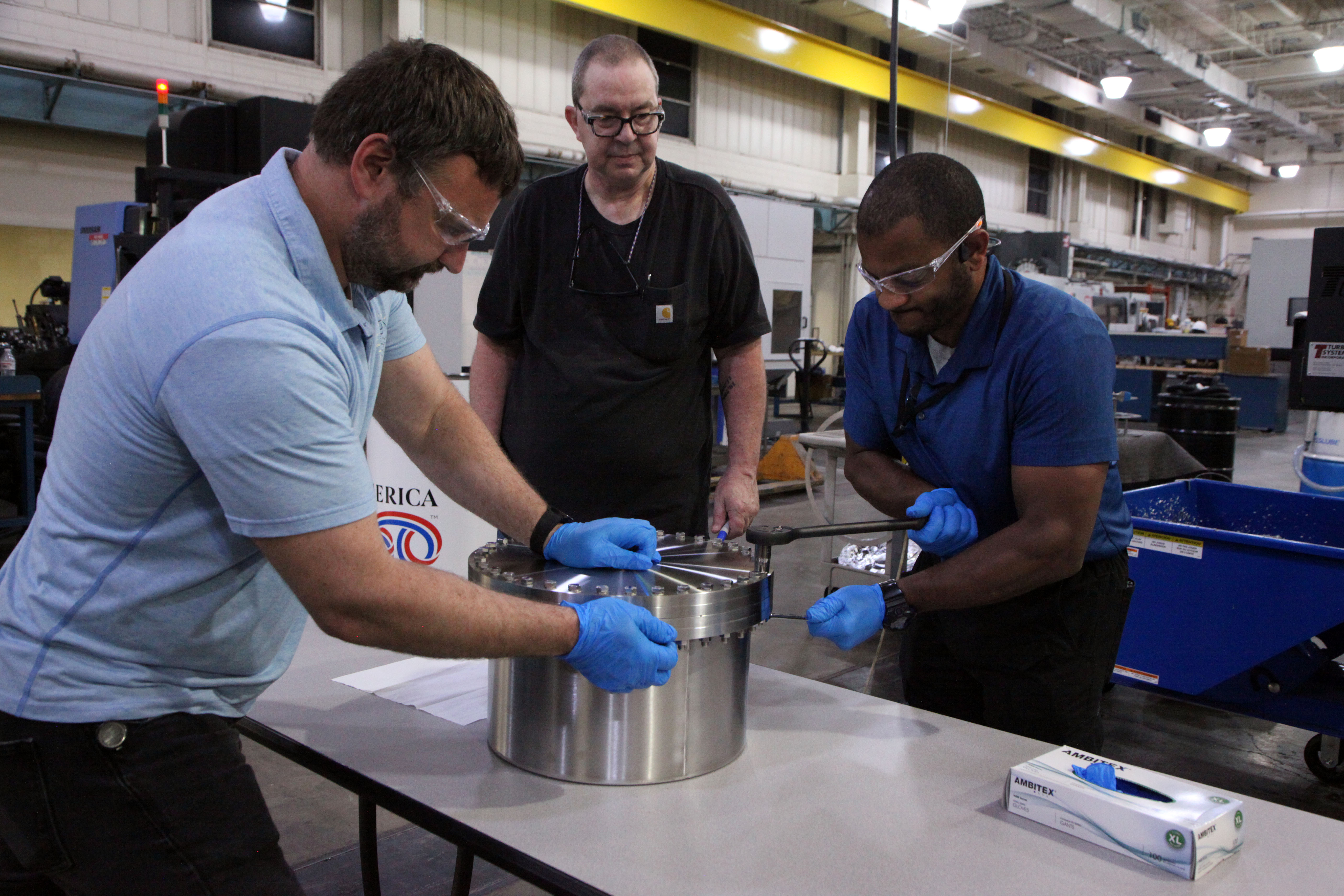 Two men are tightening a lid on a short metal cylinder in a machine shop while a third looks on.