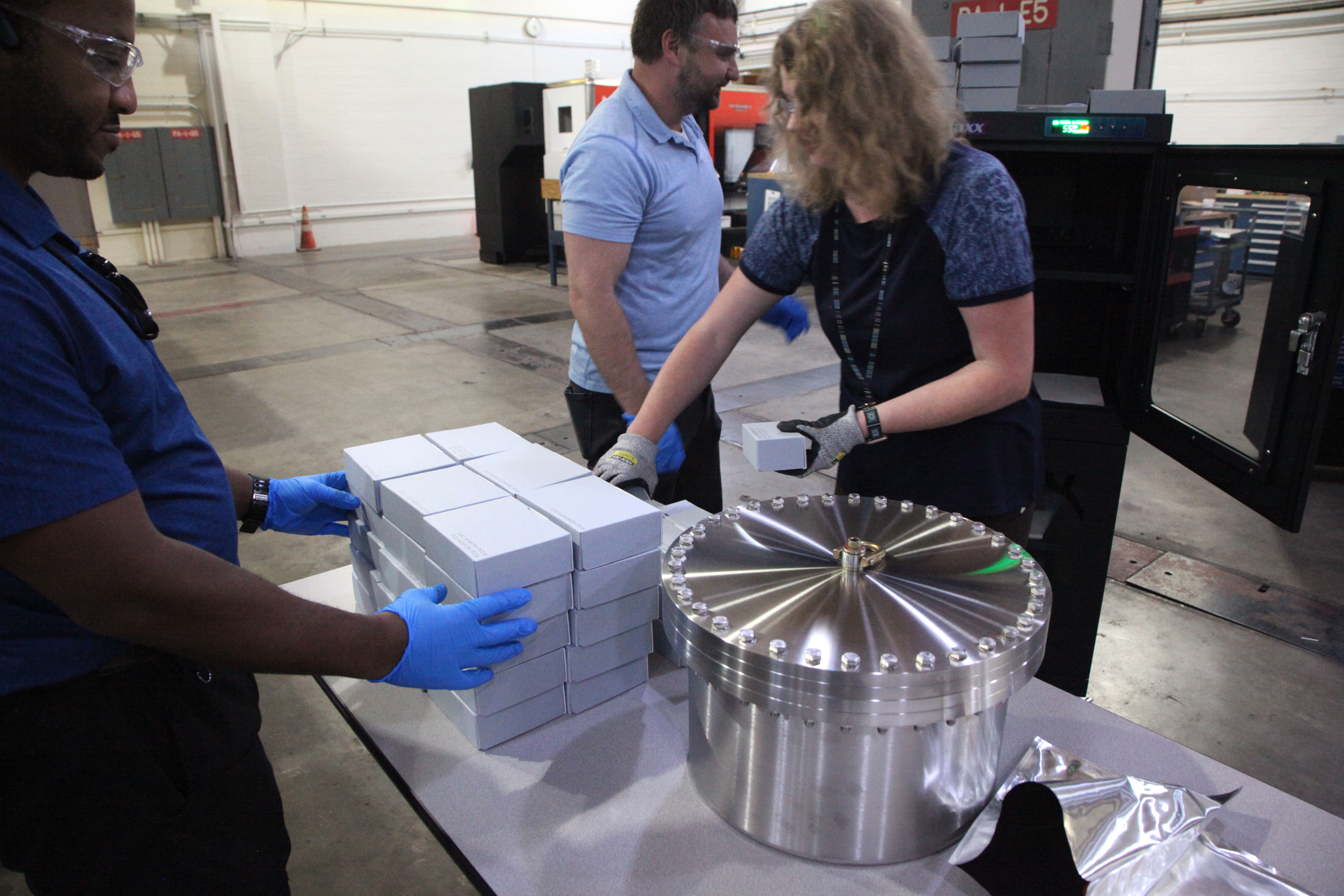 A woman wearing gloves stacks identical small boxes on a table in a machine shop, with other workers around her to help.