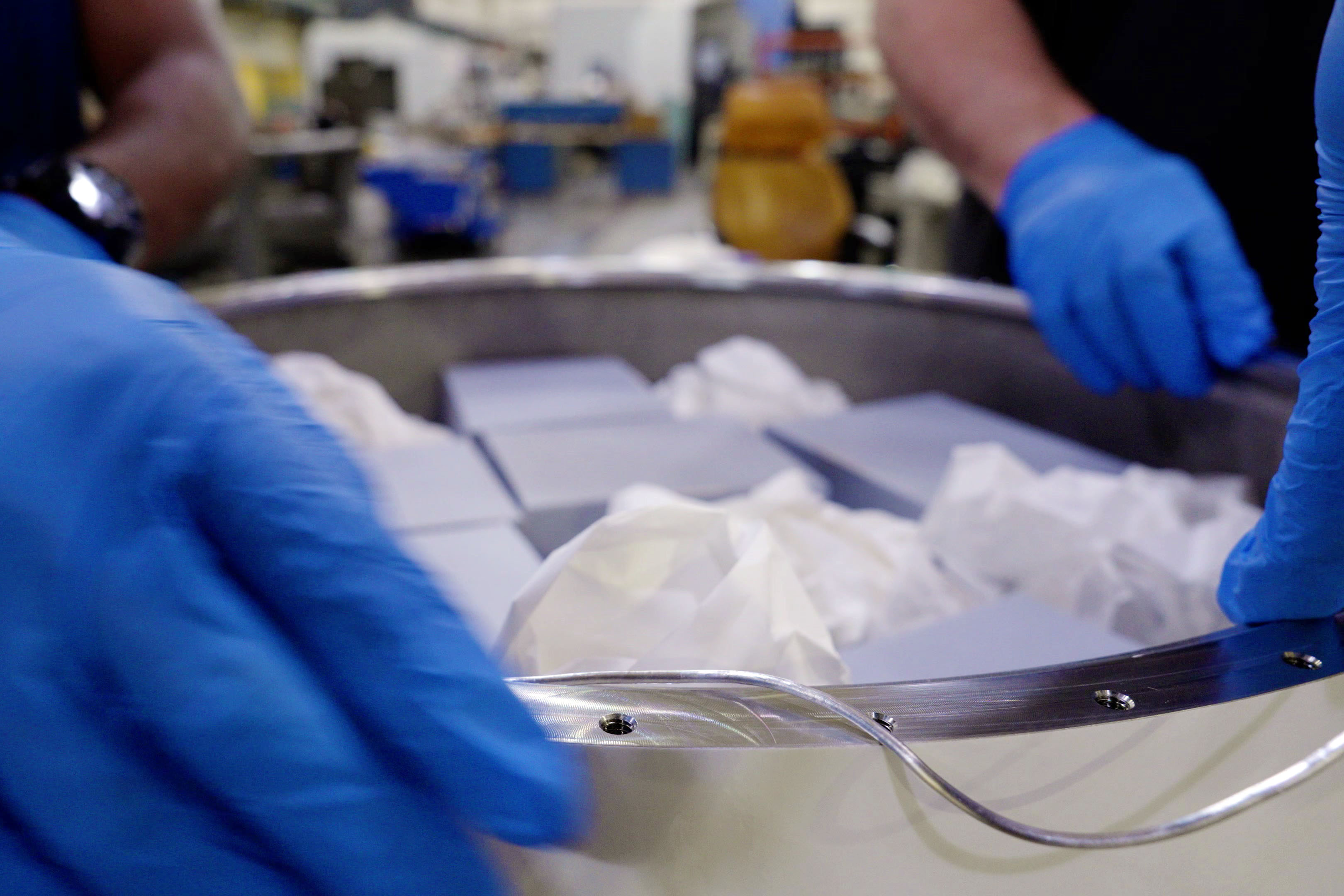 People wearing blue gloves are pressing a silver-colored wire around the rim of a circular container holding small boxes and packing material. 