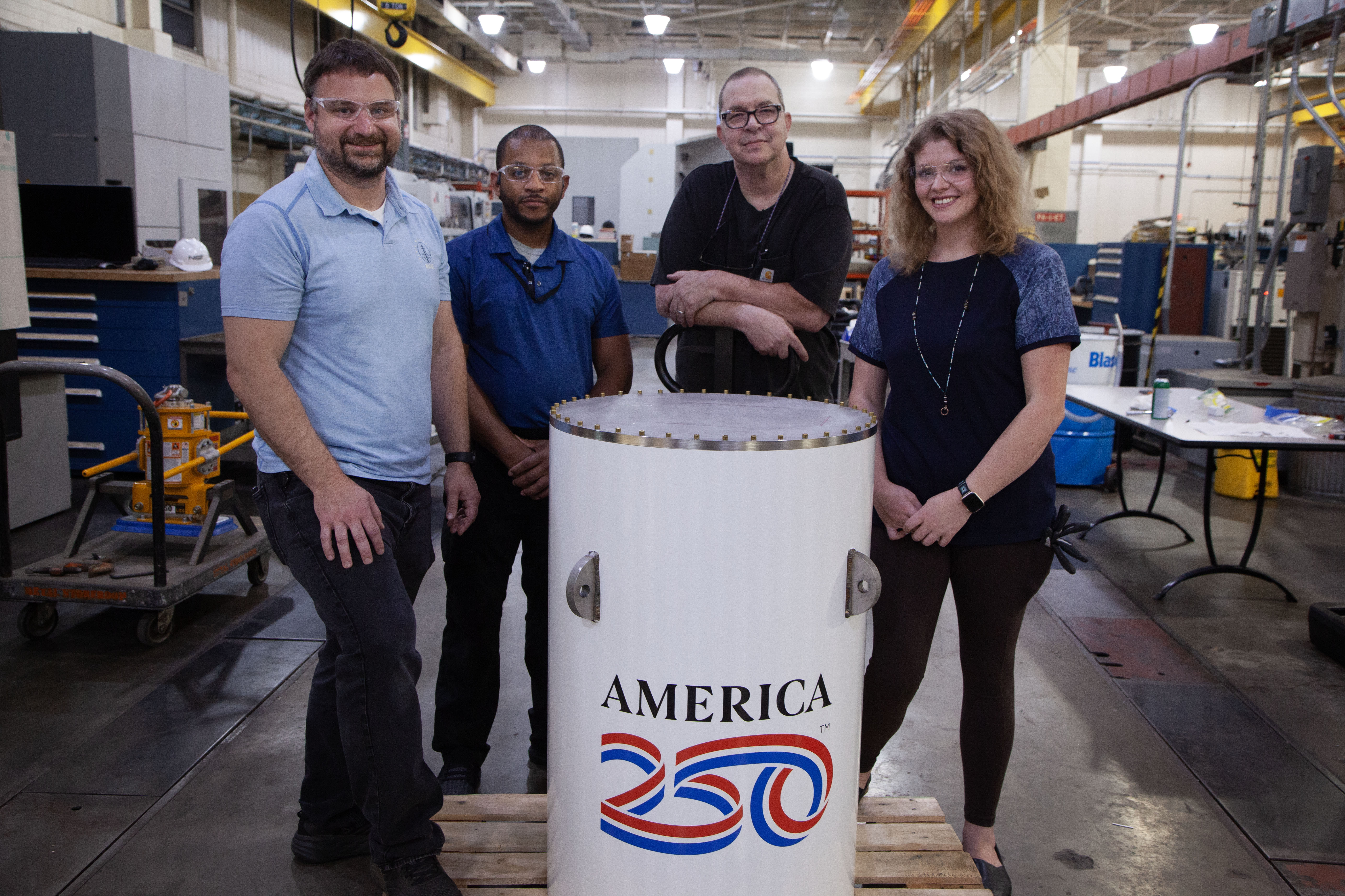 Four workers in a machine shop pose smiling around the upright white cylinder of the America250 time capsule. 