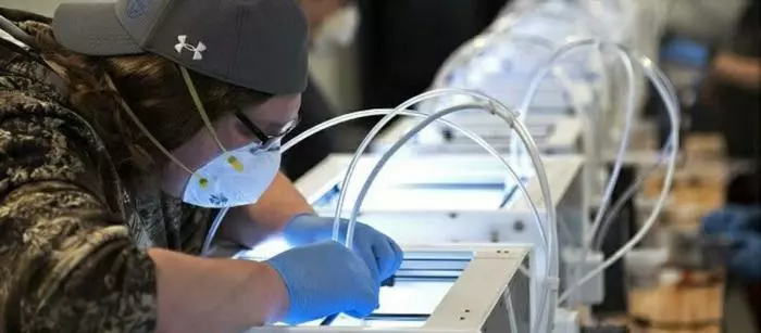 A person in a baseball cap and mask bends over a high-tech manufacturing workstation.