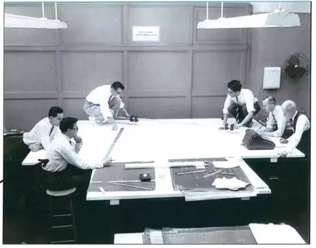 Black-and-white photo shows six men in office attire around a large table with construction plans laid out on it. 