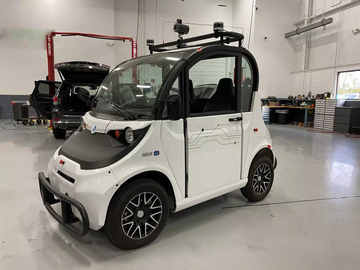 A tiny white car sits inside a large, clean garage space with tools and another vehicle in the background. 