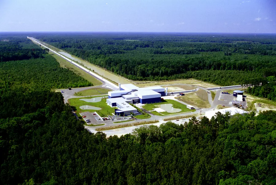 Aerial view of LIGO facility shows a white-roofed lab building surrounded by forest, with long thin paths extending from the building's back and side into the distance. 