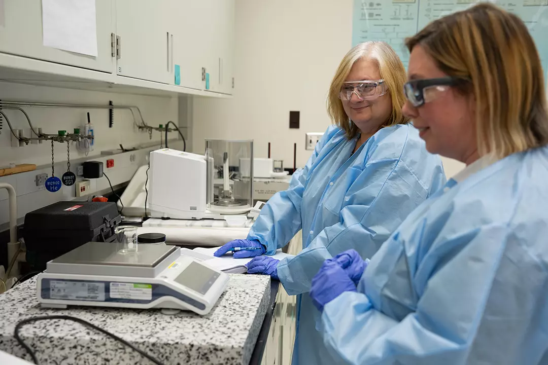 Two women in blue paper lab coats and safety glasses stand at a lab counter, looking at a small glass container on a scale. 