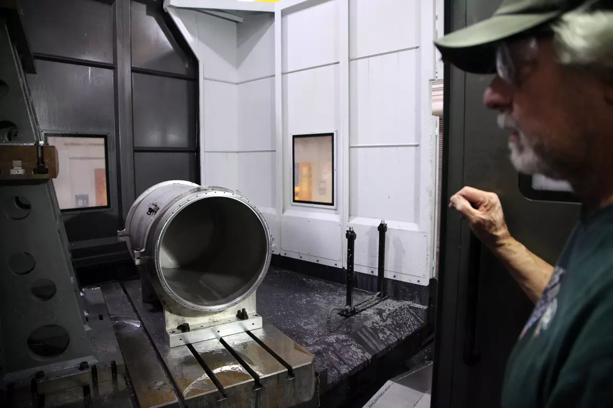 A man wearing safety glasses stands looking at a cylinder of thick metal lying on a wet platform inside a workshop. 