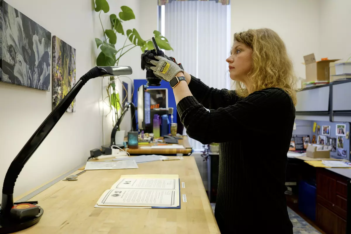 A woman wearing gloves stands in an office holding a camera pointed down at papers on the desk.