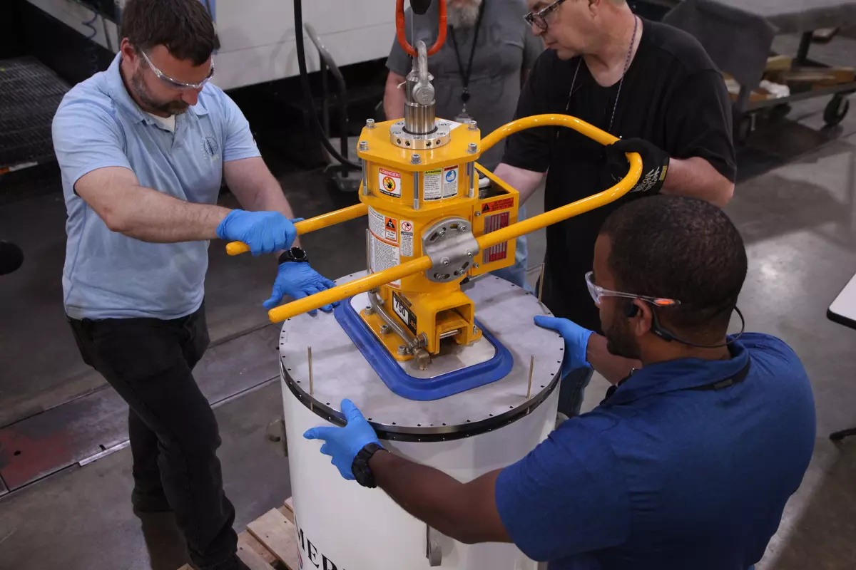 Three men stand around the cylindrical time capsule, handling a yellow device that holds the lid of the cylinder.