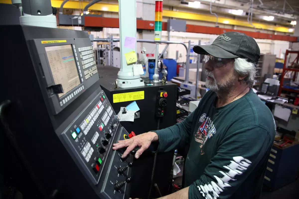 A man in a baseball cap stands at a control panel full of buttons inside a machine shop.