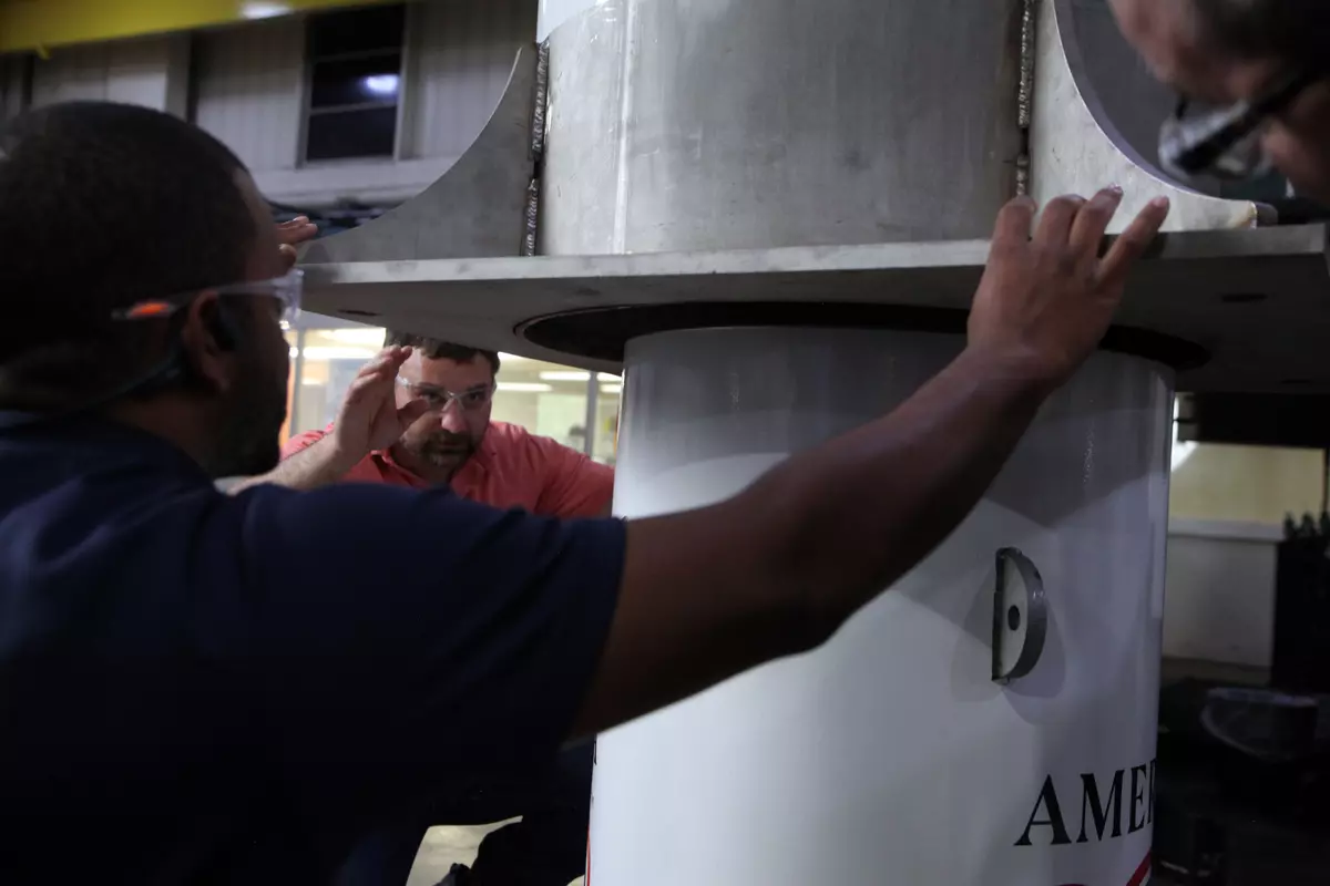 Two men reach out to hold a large metal cylinder being lowered onto a smaller one. 