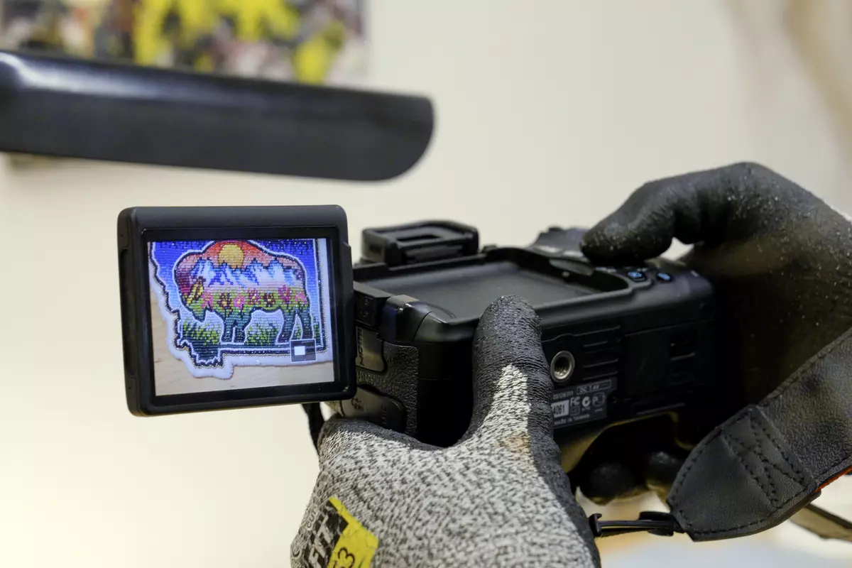 Gloved hands hold a camera; an attached screen shows a colorful beaded patch depicting a buffalo. 