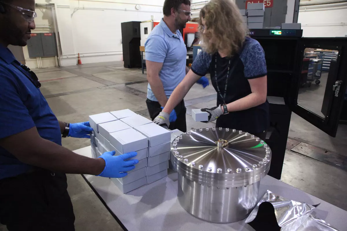A woman wearing gloves stacks identical small boxes on a table in a machine shop, with other workers around her to help.