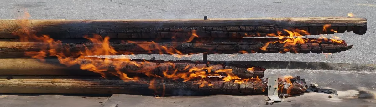 A photo of burning landscape timbers. The timbers are a stack of four rounded nominal three-by-four dimensional treated pine landscape timbers. The photo shows fire progressing along the outside and between the timbers.