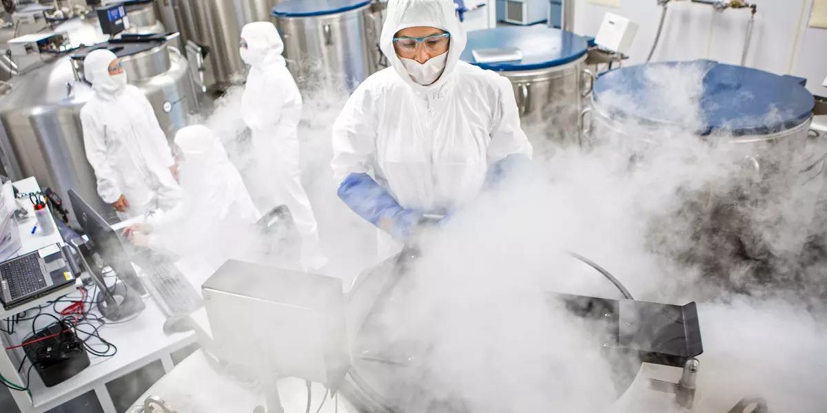 Giant metal tubs are being opened and white vapor is puffing up into the face of one researcher who is in a protective suit with hood, gloves and eye protection. In the background, several other suited researchers discuss something.