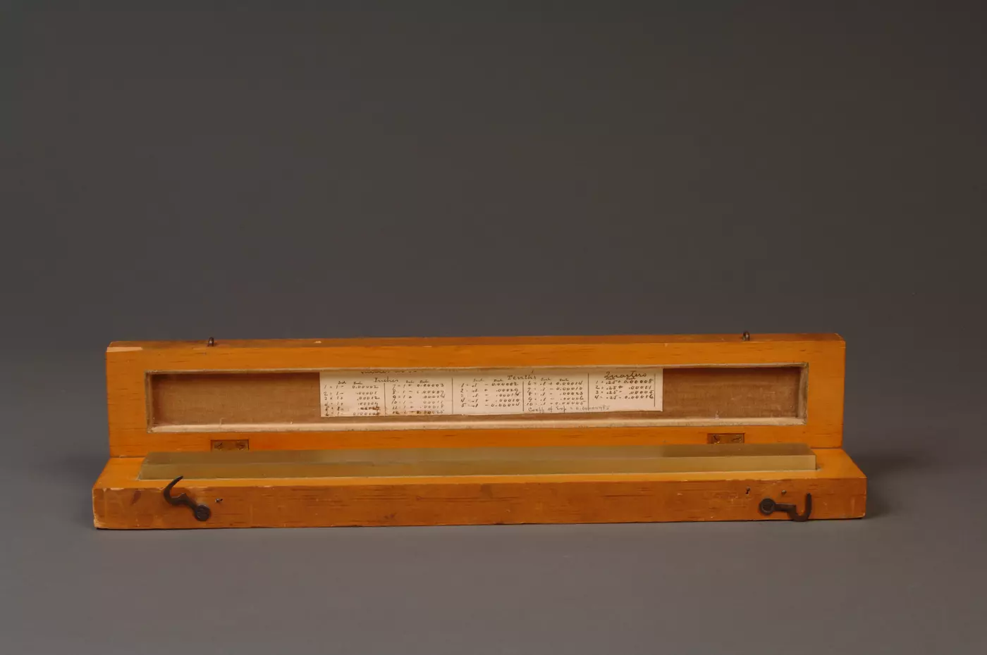Brass line scale sitting in wooden box with lid open, calibration writing visible on side of lid.