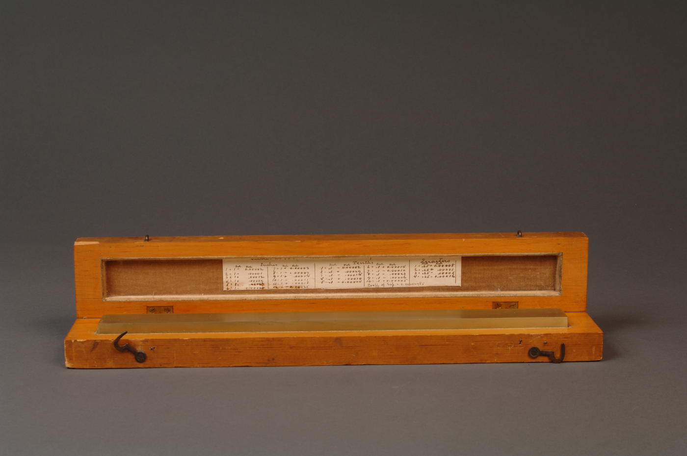 Brass line scale sitting in wooden box with lid open, calibration writing visible on side of lid.