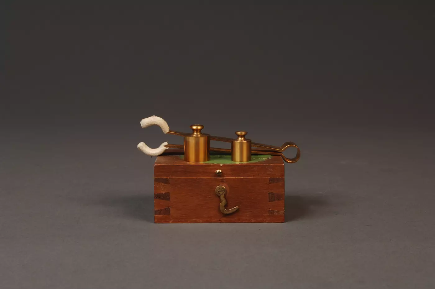 Two gold-plated brass weights sitting on top of a wood box, in front of tongs used to handle them