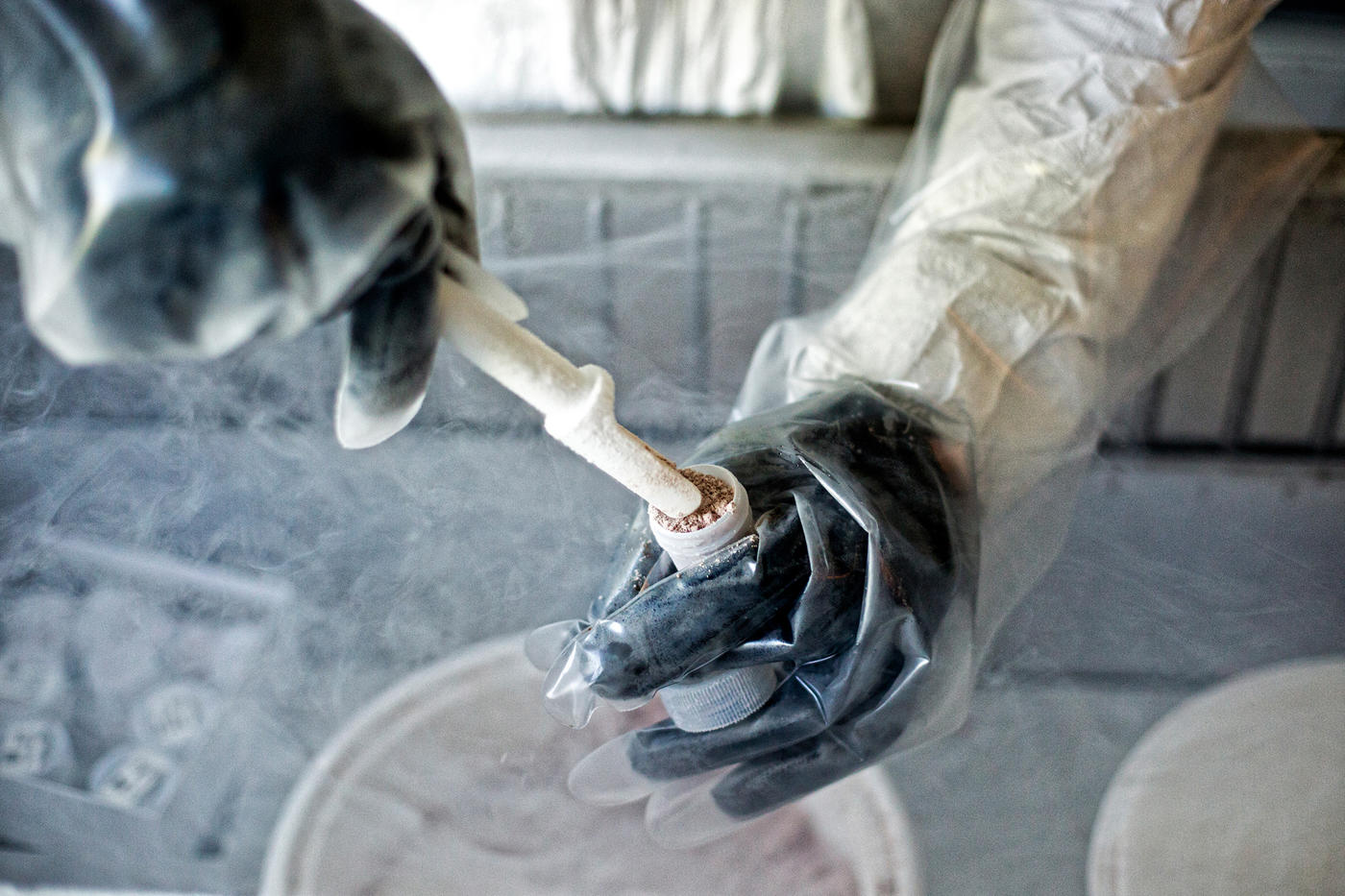 A pair of gloved hands prepares a specimen for storage using a test tube and a special, slim scoop.