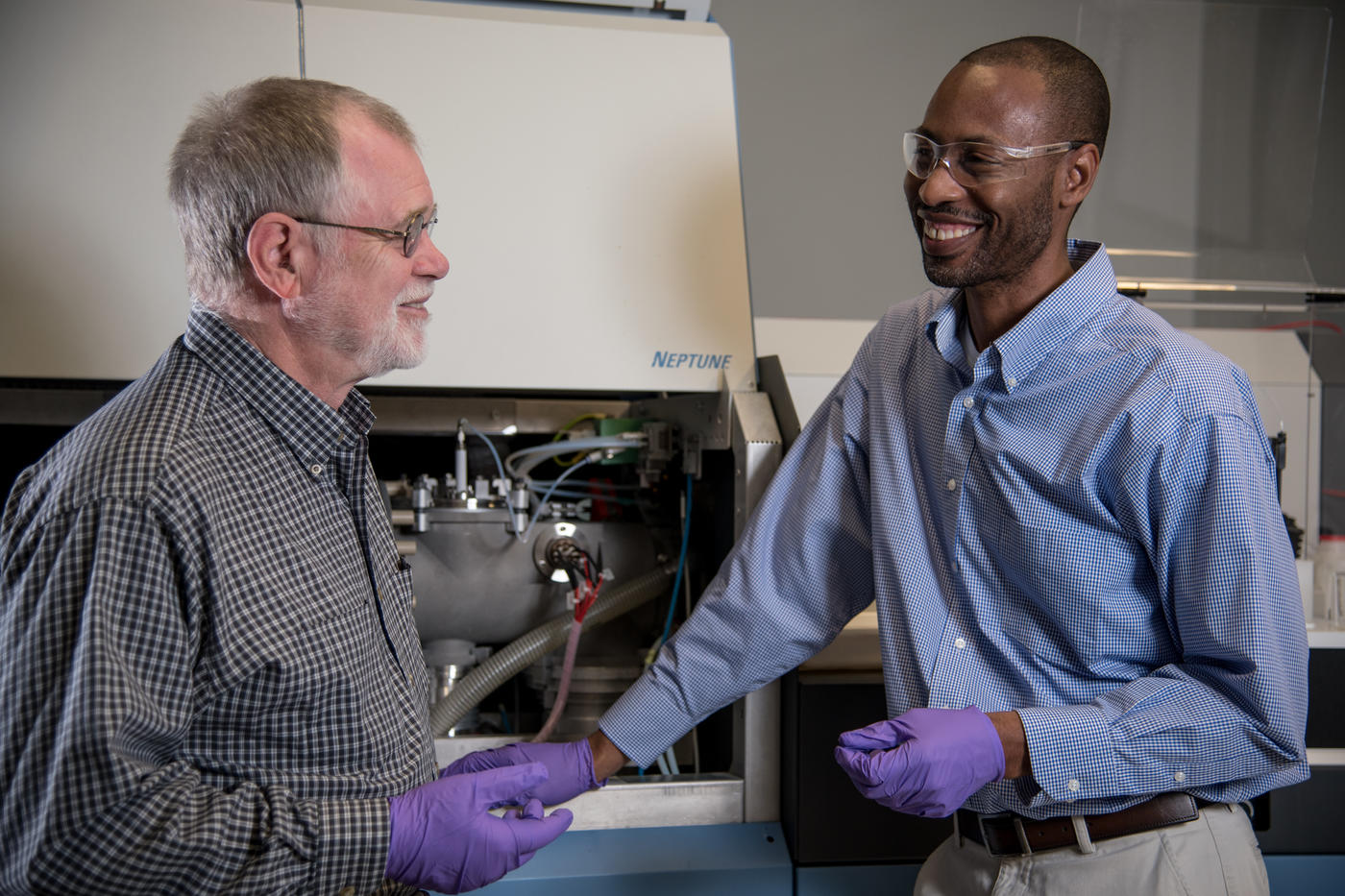 Two men (Bob Vocke and Savelas Rabb )stand in a lab setting, smiling and discussion something in front of their mass spectrometer, a large instrument used in chemistry.