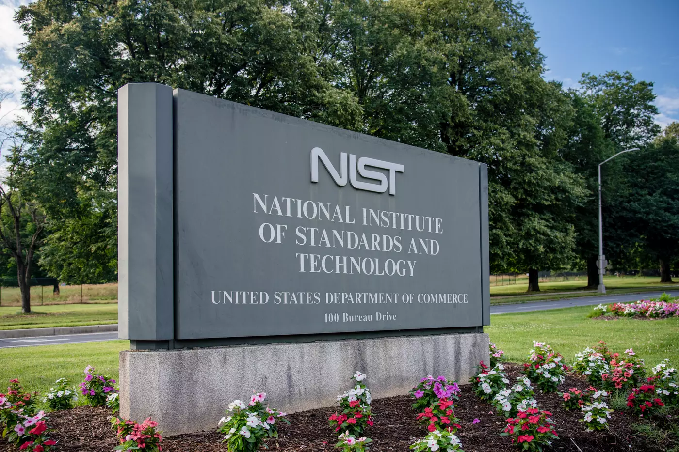 gray entrance sign that says NIST National Institute of Standards and Technology, United States Department of Commerce, 100 Bureau Drive. Surrounded by green grass, flowers, trees.