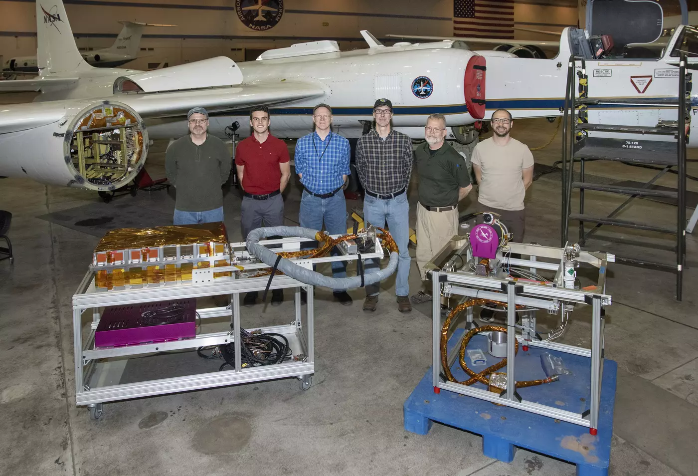 Six men stand in front of an airplane in a hangar, with equipment displayed on carts in front of them. 