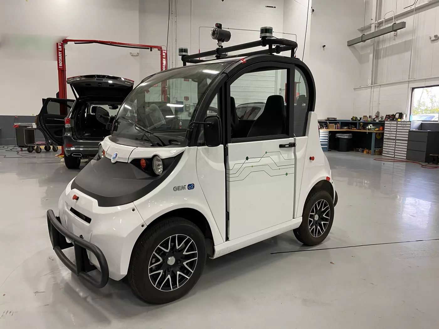 A tiny white car sits inside a large, clean garage space with tools and another vehicle in the background. 