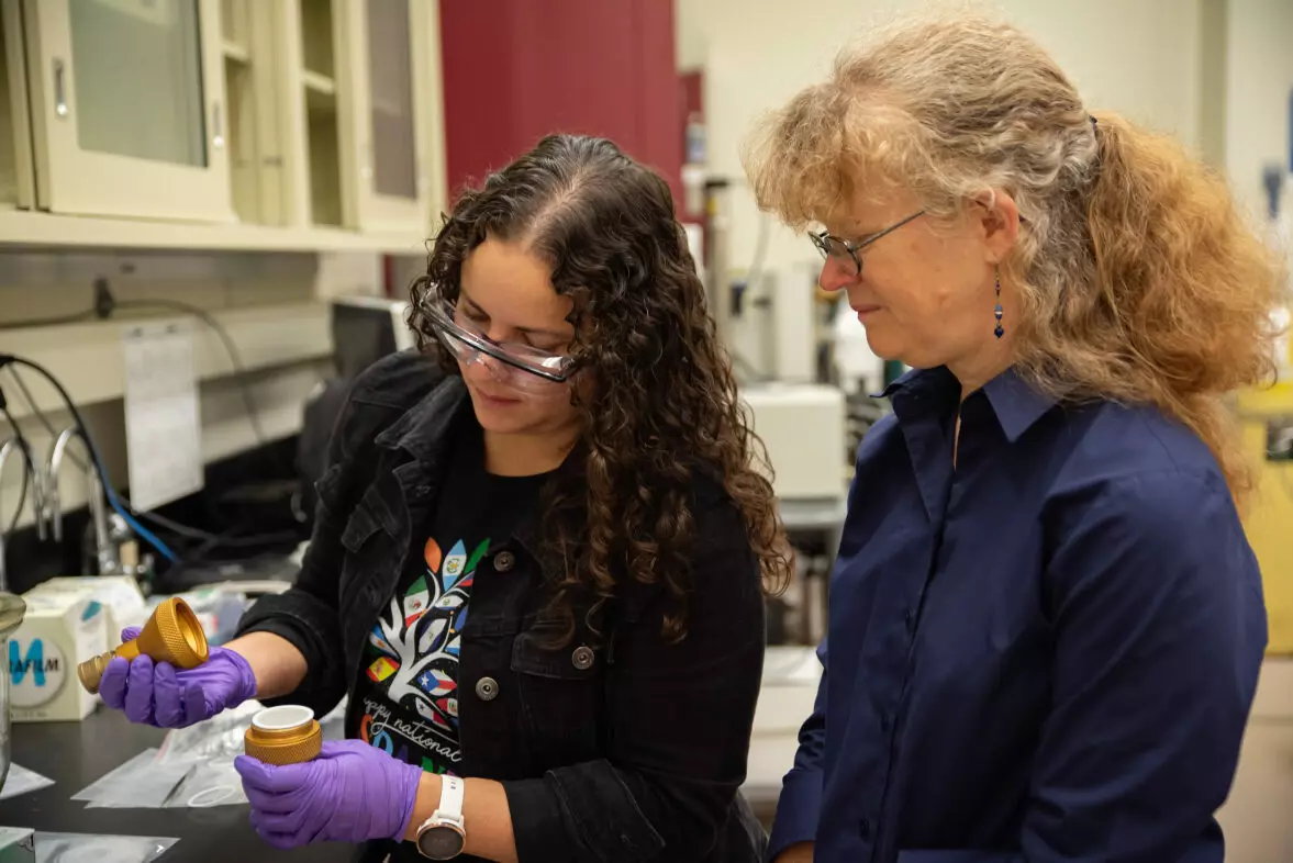 Diana Ortiz-Montalvo looks into a small container in the lab while Abigail Lindstrom stands behind her. 